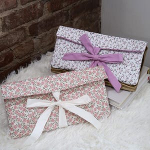 May include: Two floral print clutch bags with bows. One bag has a white bow, the other a lavender bow. The bags are white with pink flowers and green leaves. The bags are on a white fluffy surface.