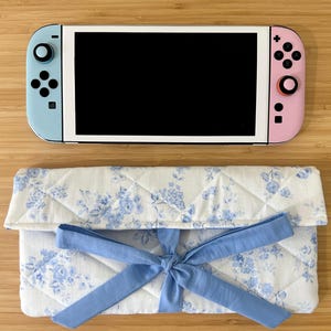 Pode incluir: Uma consola Nintendo Switch com comandos azuis e rosa repousa sobre uma capa acolchoada com padrão floral. A capa é branca com desenhos florais azuis e um laço azul claro. O ecrã da consola é preto.