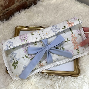 Pode incluir: Uma capa acolchoada com estampa floral e laço azul claro. A capa apresenta um fundo branco com um padrão de flores rosa, roxas e azuis. As bordas são adornadas com renda branca. A capa está sobre uma moldura dourada.