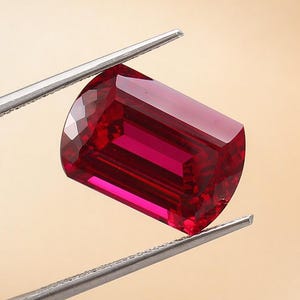 May include: A faceted, rectangular ruby gemstone held by silver tweezers. The ruby is a deep, saturated red color with a polished surface. The background is a soft, warm beige.