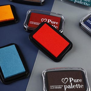 Könnte beinhalten: Eine Sammlung farbiger Stempelkissen mit schwarzen Basen und transparenten Deckeln. Die Kissen haben leuchtende Farben wie Rot, Orange und Blau. Einige Kissen tragen den Text "Pure palette" und "GLAY SHADES".