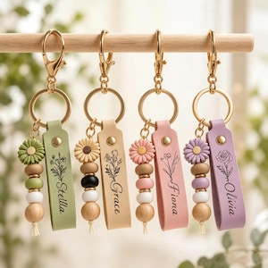Pode incluir: Quatro porta-chaves com ferragens douradas, cada um com uma tira de couro com um desenho floral e um nome: Stella, Grace, Fiona ou Olivia. Cada um inclui um pendente em forma de flor e três contas de madeira em cores coordenadas.