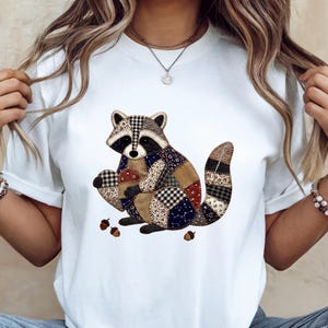 May include: A white short-sleeved t-shirt featuring a central graphic of a raccoon designed with a patchwork pattern. The raccoon illustration incorporates various fabric textures, including plaid, floral, and solid brown patches, while holding small acorns.