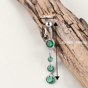 Puede incluir: Un piercing de ombligo plateado con un diseño de barra curva. Cuenta con una bola superior, una gema verde grande y tres gemas verdes colgantes en forma de lágrima. La longitud total es de 4,9 cm.