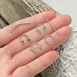 Puede incluir: Una colección de joyas corporales plateadas, que incluye barras curvas con piedras preciosas de varios colores y una barra recta. Las piedras preciosas son de color azul, verde, rosa y transparente. También se incluye una aguja curva.