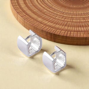 May include: A pair of silver-colored, square-shaped hoop earrings. The earrings are displayed on a light tan surface, with a wooden, circular object in the background. The earrings have a polished, metallic finish.
