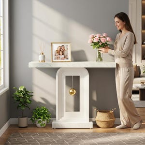 May include: A white console table with a marble-patterned top and base, featuring an arched support structure with a suspended gold sphere. The table holds a gold-framed photograph of a family and a clear glass vase filled with pink roses.