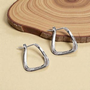 May include: A pair of silver-toned, geometric hoop earrings. The earrings have a hammered texture and a triangular shape. They are displayed on a light tan surface with a wooden slice in the background.