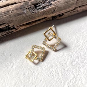 May include: A pair of gold-toned stud earrings. Each earring features two connected square shapes, one slightly smaller than the other. The outer edges of the squares are lined with small, clear crystals, adding sparkle to the jewelry.