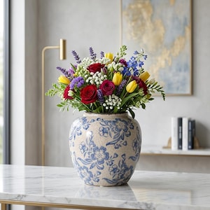 May include: A round ceramic vase with a cream-colored base and blue floral patterns featuring a crackled glaze finish. The vase is filled with a vibrant bouquet of red roses, yellow tulips, purple lavender, blue delphiniums, and white baby's breath.
