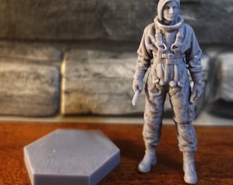 May include: A grey 3D printed miniature figurine of a person wearing a flight suit with a harness and a collar, standing next to a separate grey hexagonal base. The figure has short hair and is holding a small cylindrical object in one hand.