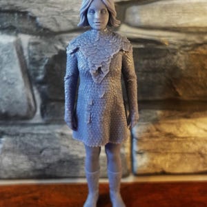 May include: A monochromatic gray 3D printed figurine of a humanoid character with antennae, short hair, and a textured, scale-like outfit. The figure wears a dress with a layered, petal-like collar and matching boots, standing in a neutral, upright pose.
