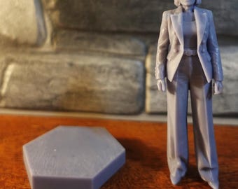 May include: A light grey 3D printed miniature figurine of a person wearing a double-breasted blazer and wide-leg trousers, standing next to a separate light grey hexagonal base. The figure has short hair and a formal, structured appearance.