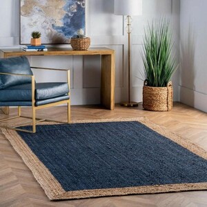 May include: A rectangular navy blue rug with a natural fiber border. The rug is placed on a hardwood floor, and is surrounded by furniture and decor, including a blue chair, a wooden table, and a tall plant.