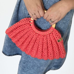 May include: Coral-coloured, hand-knitted handbag with a wooden handle. The bag features a ribbed texture and a small, circular wooden tag. The handle is a light brown colour. The bag is held by a person wearing a denim dress.