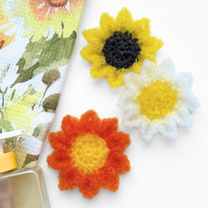 Op de afbeelding: Drie bloemvormige schuursponsjes met een textuur. Eén heeft gele blaadjes en een zwart hart, één heeft witte blaadjes en een geel hart, en één heeft oranje blaadjes en een geel hart. Een fles handzeep en een bloemige doek liggen in de buurt.