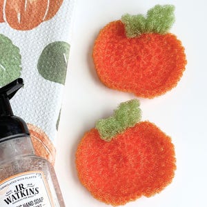 May include: Two round, orange, crocheted pumpkin-shaped scrubbers with green stems. Beside them is a clear plastic bottle of J.R. Watkins foaming hand soap with a white label featuring orange illustrations and text, and a white towel with pumpkin prints.