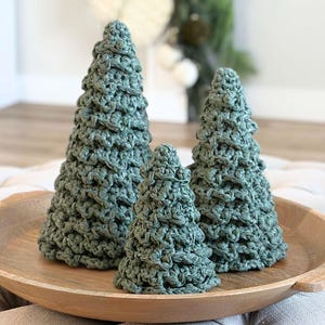 May include: Three cone-shaped decorative objects crafted from sage green yarn in a textured crochet stitch pattern. The items are arranged in a cluster, with two taller pieces flanking a smaller central piece, all resting on a round wooden tray.