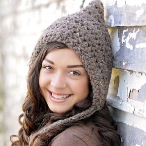 May include: A brown, chunky knit beanie with a pointed top and ear flaps. The hat is crafted from a textured yarn featuring subtle flecks of lighter color throughout the weave, providing a thick, cozy appearance for cold weather headwear.
