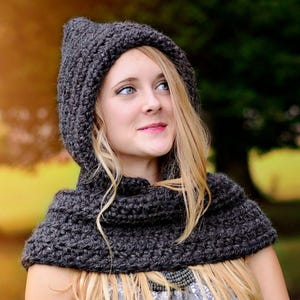 May include: A dark charcoal gray hooded cowl made of thick, chunky knit yarn. The accessory features a pointed hood design that transitions into a wide, draped neck warmer that sits over the shoulders, showcasing a textured, ribbed stitch pattern.