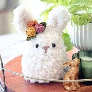 May include: A fluffy white bunny plush toy with a floral crown, a small gold bunny figurine, and a green candle in a decorative holder sit on a wooden tray. A white potted plant is in the background.