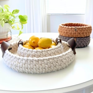 May include: A set of three handmade crochet baskets in varying sizes. The largest basket, filled with lemons, has leather handles. The other two baskets are in shades of gray and orange. The baskets are on a white table.
