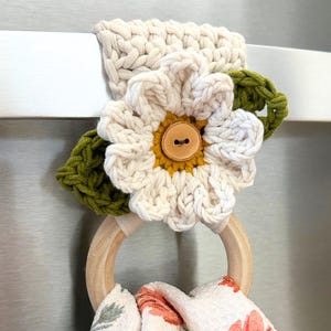 Puede incluir: Un portatoallas de ganchillo hecho a mano con una gran flor blanca con centro amarillo y un botón de madera. El soporte incluye un anillo de madera y una sección superior tejida de color crema. Una toalla con estampado floral cuelga del anillo.