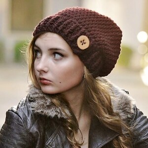 Puede incluir: Un gorro de ganchillo color granate oscuro con un botón de madera claro en el lateral, combinado con una chaqueta de piel sintética negra con cuello de pelo sintético, una camiseta blanca, vaqueros azules y botines marrones con cordones.