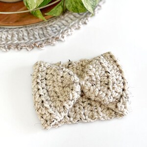May include: A cream-colored crochet headband featuring a thick, chunky yarn texture with subtle black flecks throughout. The design includes a central knotted or twisted detail that creates a gathered effect, showcasing a dense, hand-stitched pattern.