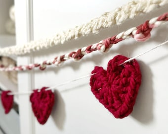 May include: A decorative garland featuring a series of small, dark red crocheted hearts strung along a thin white cord, positioned below two additional strands of textured, twisted yarn in cream and variegated red and white tones.
