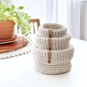 May include: A stack of four round, off-white crocheted baskets of varying sizes. Each basket features a vertical tan leather tag attached with a metal rivet, with the word "smbe" embossed in cursive script on each tag.