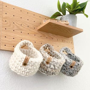 May include: Three small, round, crocheted hanging baskets in cream, beige, and grey yarn. Each basket features a tan leather tag with the text "smbe" attached by a brass-colored metal rivet. The baskets are suspended from a light wood pegboard.