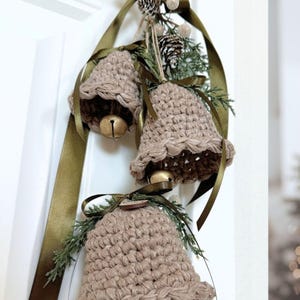 May include: Three crocheted beige bells hanging from a dark olive green satin ribbon. Each bell features a metallic gold jingle bell inside and is decorated with small green pine sprigs, a tiny pinecone, and a small wooden circular accent.