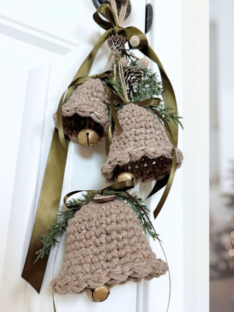 May include: Three crocheted beige bells hanging from a dark olive green satin ribbon. Each bell features a metallic gold jingle bell inside and is decorated with small green pine sprigs, a tiny pinecone, and a small wooden circular accent.
