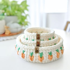 May include: A set of three nested, round, cream-colored crocheted baskets featuring a repeating pattern of small orange carrots with green tops. Each basket has a small, vertical, tan-colored leather-like tag attached to the front rim.