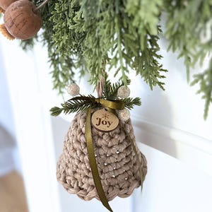 May include: A beige crocheted bell ornament featuring a dark olive green ribbon tied in a bow at the top. A small circular wooden tag is attached to the ribbon, engraved with the word Joy and a star symbol. The bell is accented with small sprigs of faux pine needles and white berries.