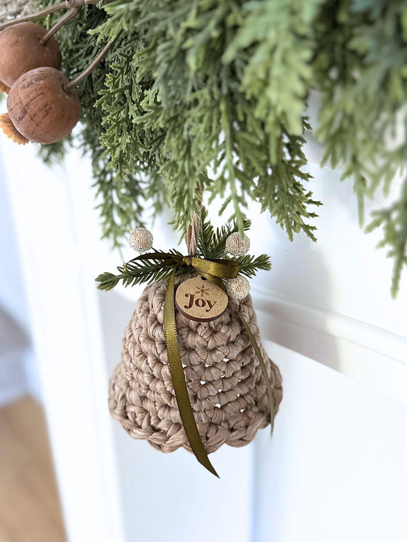 May include: A beige crocheted bell ornament featuring a dark olive green ribbon tied in a bow at the top. A small circular wooden tag is attached to the ribbon, engraved with the word Joy and a star symbol. The bell is accented with small sprigs of faux pine needles and white berries.