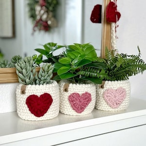 Op de afbeelding: Drie crèmekleurige gehaakte plantenpotovertrekken, elk voorzien van een hartapplicatie in rood, donkerroze of lichtroze. Elke hoes heeft een klein bruin lederachtig label met het woord LOVE in reliëf op de voorzijde.