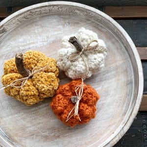 May include: Three crocheted pumpkins in mustard yellow, cream white, and burnt orange. Each pumpkin features a textured yarn surface, a natural wooden twig stem, and a small decorative ribbon tied around the stem base for a rustic appearance.