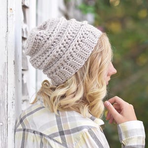Könnte beinhalten: Eine hellbeige, grob gestrickte Mütze mit einem strukturierten Rippenmuster und vertikalen Maschen. Die Mütze hat eine lockere, abgerundete Form, die den Kopf bedeckt, und ist aus dickem Garn in einer einheitlichen, neutralen Farbe gefertigt.
