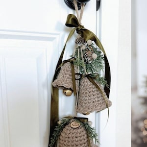 May include: Three crocheted beige bells hanging from a dark olive green ribbon. Each bell is adorned with green pine sprigs, small pinecones, and a wooden tag with the word Noel engraved on it. Gold jingle bells are visible inside the crochet.
