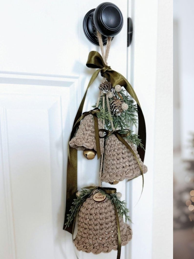 May include: Three crocheted beige bells hanging from a dark olive green ribbon. Each bell is adorned with green pine sprigs, small pinecones, and a wooden tag with the word Noel engraved on it. Gold jingle bells are visible inside the crochet.