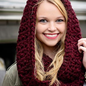 May include: A chunky, deep burgundy knit infinity scarf worn as a hood. The scarf features a thick, textured yarn construction with a prominent braided stitch pattern that frames the face, providing a warm and cozy appearance in a dark red hue.