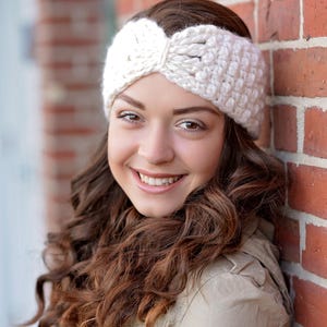 May include: A thick, off-white crocheted headband featuring a central twisted knot detail. The accessory is crafted from chunky yarn with a visible, textured stitch pattern that wraps around the head, providing a wide band coverage.