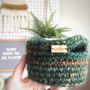Peut inclure: Un panier rond crocheté à la main en fil panaché dans des tons de vert forêt, marron et des touches d'or. Une petite étiquette rectangulaire en bois est fixée sur le devant, portant le texte gravé "smbe" avec une petite icône de cœur.
