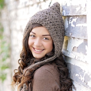 May include: A brown, chunky knit beanie with a pointed top and ear flaps. The hat is crafted from a textured, speckled yarn in a neutral earth tone, featuring a thick crochet stitch pattern that covers the head and extends down the sides.