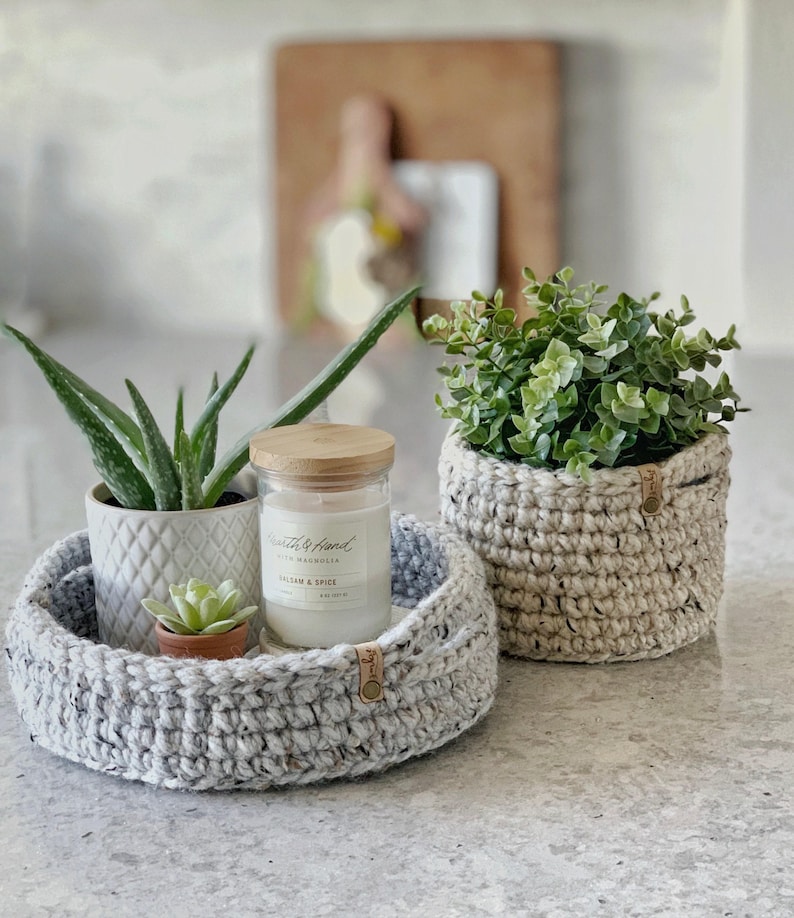 May include: A round, speckled gray crochet basket holds a small succulent, a candle in a glass jar, and a potted aloe vera plant. A second, matching crochet basket contains a green plant. The candle reads "Health & Hand, Balsam & Spice."