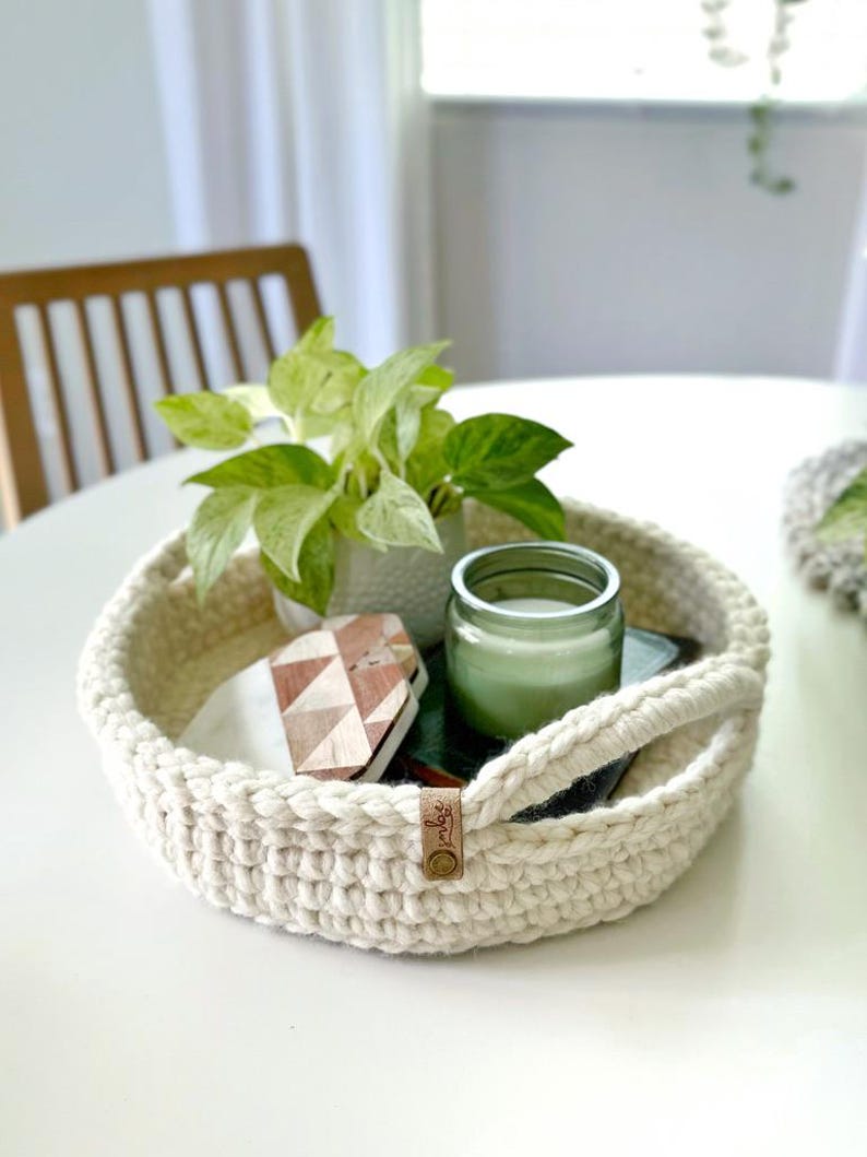 May include: A cream-colored, round crochet tray with handles, holding a small potted plant, a candle in a glass jar, and decorative items. The tray has a leather tag with a logo.
