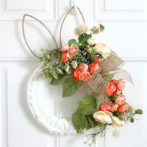 May include: A white braided wreath shaped like a bunny with two tall jute rope ears. The wreath is decorated with a cluster of peach and cream-coloured artificial roses, green leaves, small white filler flowers, and a large tan burlap ribbon bow.