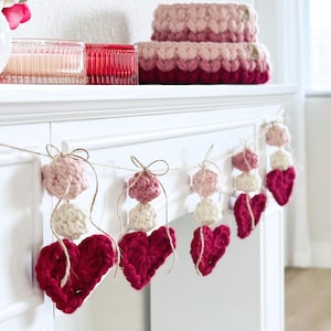 May include: A decorative garland featuring crocheted elements suspended on a thin twine string. The garland consists of repeating patterns of small light pink spheres, cream-colored spheres, and larger dark red crocheted hearts.